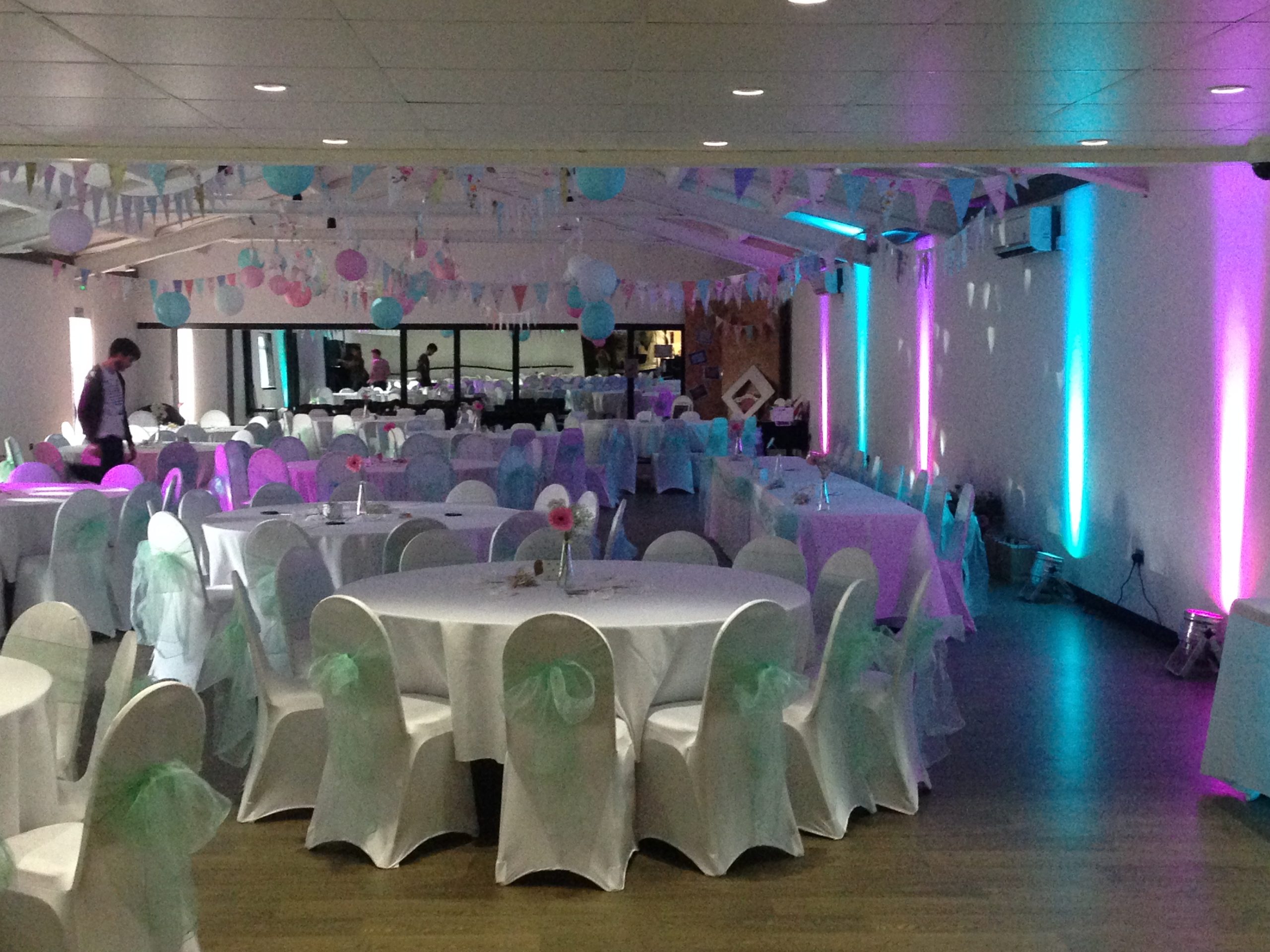 Rows of round tables with white chair covers, bunting and coloured uplighting.