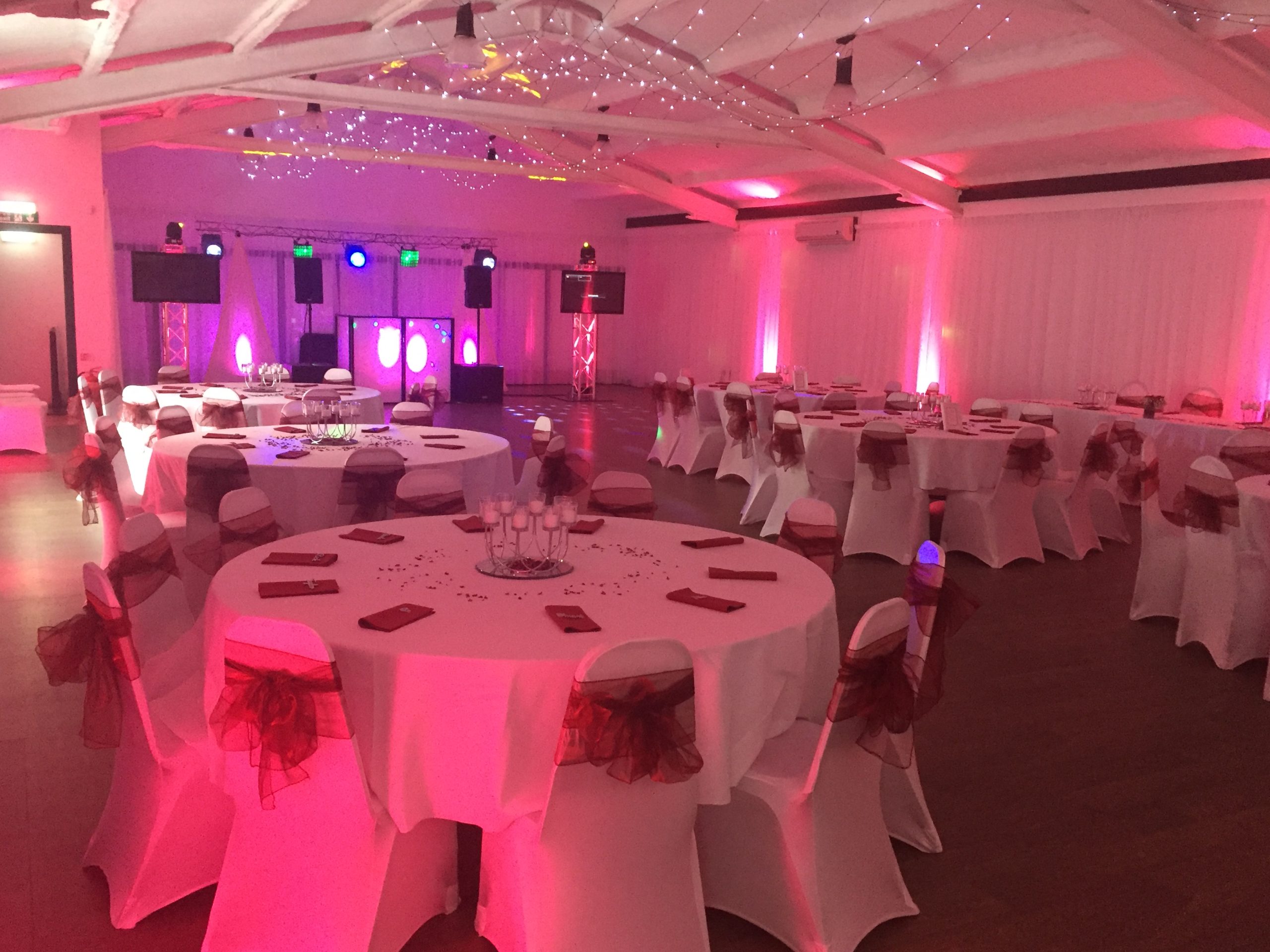 Round tables with white cloths, chair covers and burgundy sashes set for a wedding reception.